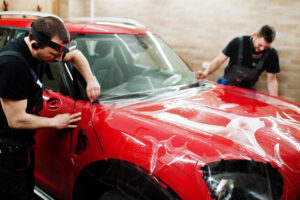 Car service worker put anti gravel film on a red car body at the detailing vehicle workshop. Car protection with special films.